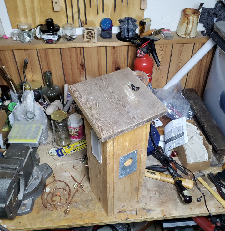 Messy workbench with birdhouse on it - the top of which has two bolts protruding through, one with a wingnut.